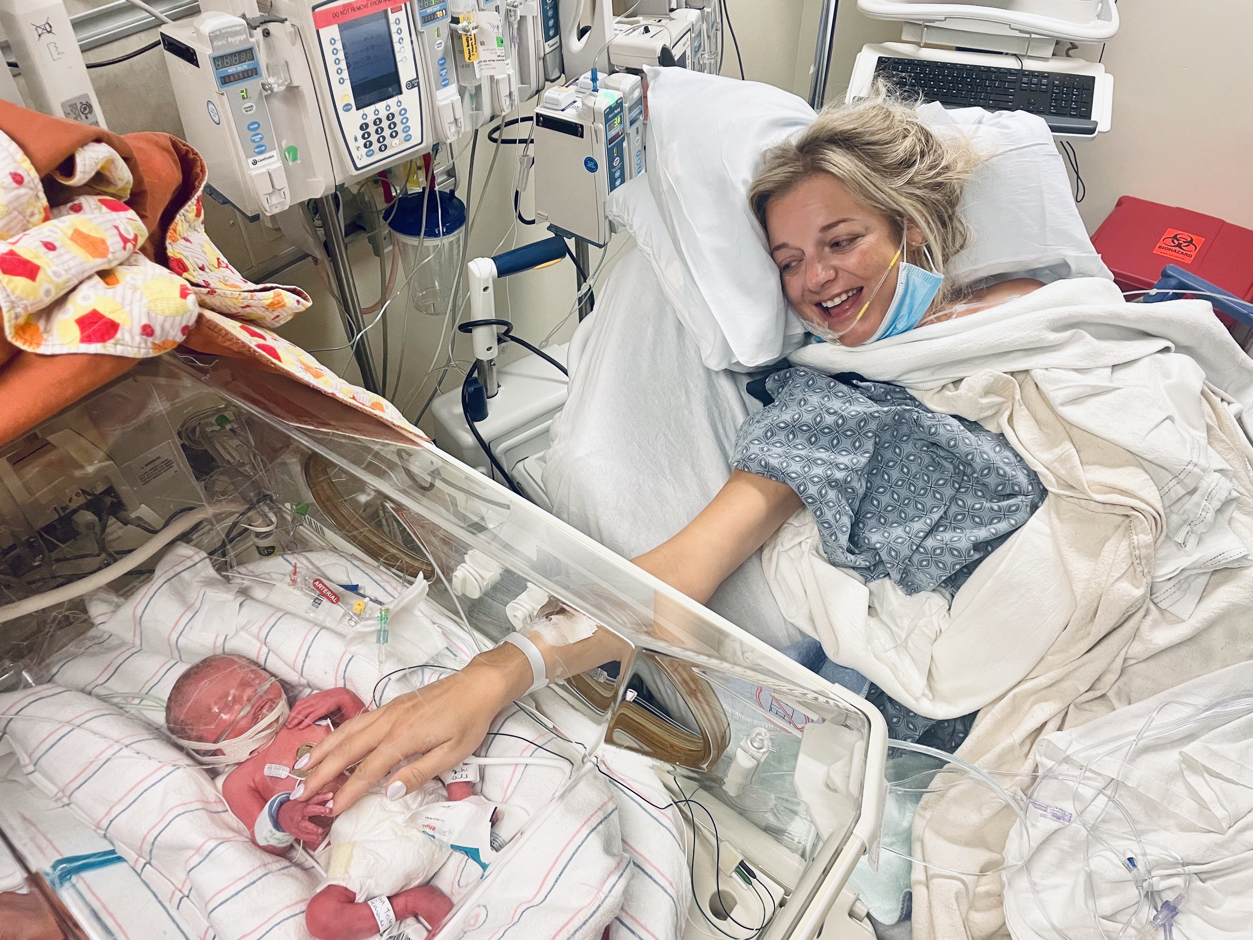 Baby Monroe lies in a NICU bed at South Shore Hospital shortly after birth, as mother Jillian reaches out to touch her, smiling, from her own hospital bed.