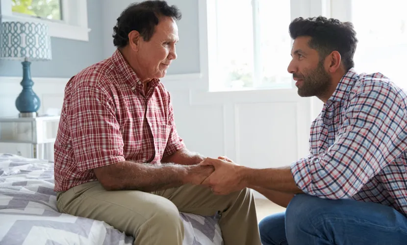 An older man smiles sitting on the edge of his bed as he talks to his adult son, who is holding his hand.