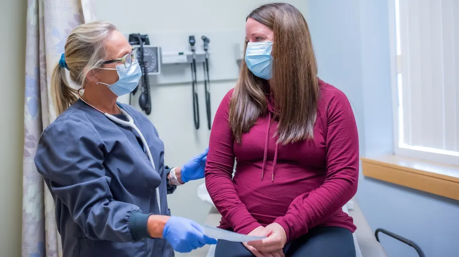 A provider at the Women's Center in Weymouth, wearing a mask and stethoscope, hands a piece of paper to a patient who is seated on an exam table.