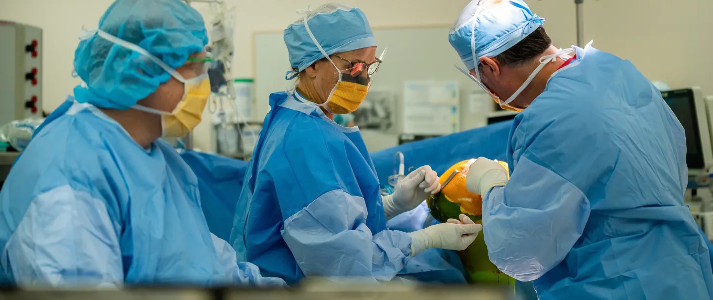 Three surgeons wearing blue scrubs, caps, and gloves crowd around a patient during a surgical procedure at South Shore Health