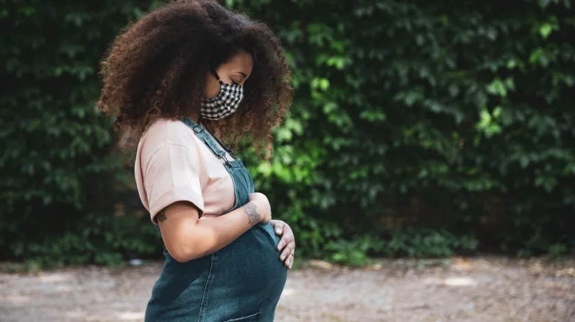 A pregnant African American woman poses outside while holding her stomach tenderly.