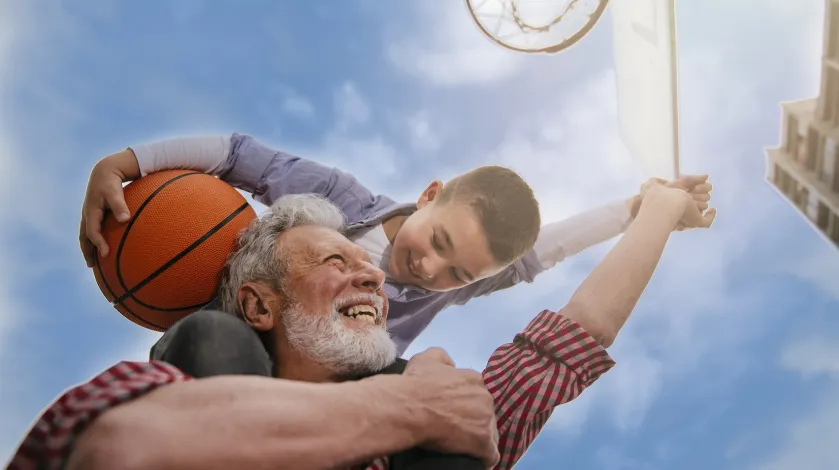 An older man smiles while playing basketball with his grandson on his shoulders
