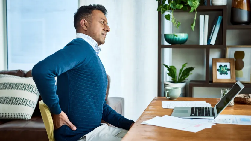 A man holds his lower back and grimaces while sitting at his dining room table for a day of remote working