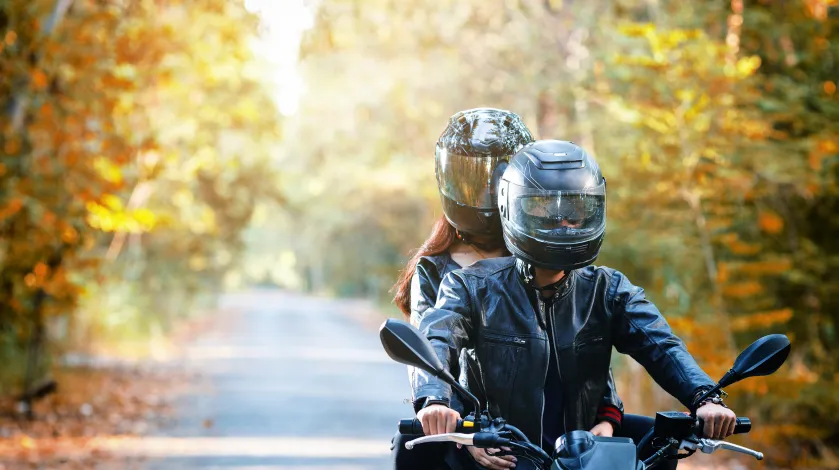 Two people wearing helmets riding on a motorcycle on a road during fall