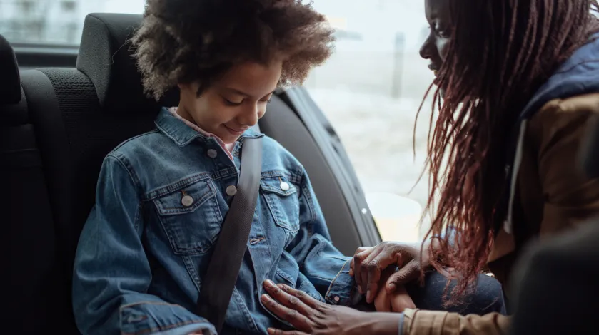 A young child looks down as her mother buckles her seatbelt in the backseat of a car.