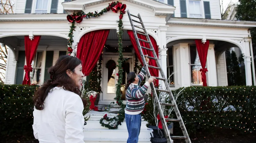 Man using a ladder to hang Christmas lights on his house