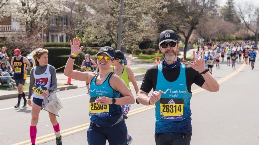 Two runners wave while making their way through the Boston Marathon route