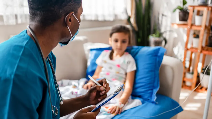 A male nurse writes on a clipboard as he speaks to a young girl patient, who is seated on a comfortable chair with an IV in her arm at her home.