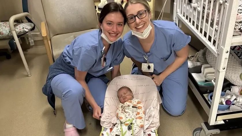 NICU baby Monroe sits in a bounce chair while two nurses from South Shore Hospital, who cared for her during her stay, smile for a photo behind her.