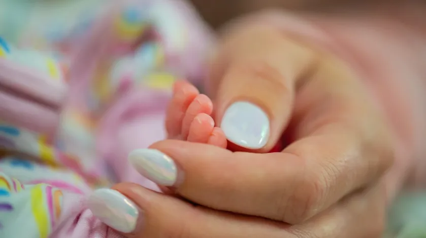 A close-up view of a mother with manicured nails gently touching the foot of her daughter, who is in a bed in the neonatal intensive care unit