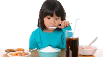 Little girl eating a spoonful of sugar from a bowl of sugar surrounded by soda and unhealthy food
