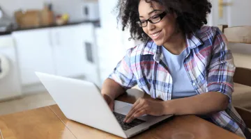 A young woman sits at her dining room table working on a white laptop, smiling while she looking at the screen