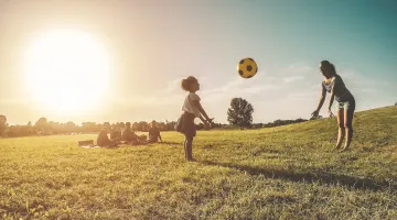 A child catches a soccer ball thrown by her mother in a field while the family has a picnic behind them. The sun is shining.