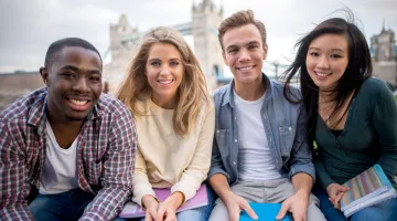 A diverse group of students holds textbooks while sitting with the Tower Bridge in London in the background
