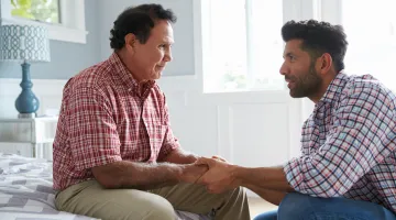 Son talks with his father who's sitting on his bed holding hands