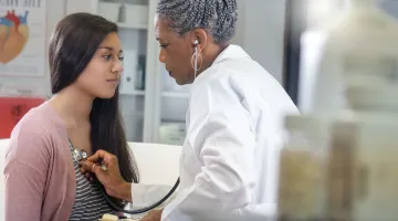 A doctor in a white coat places her stethoscope on the chest of a young girl during a physical exam