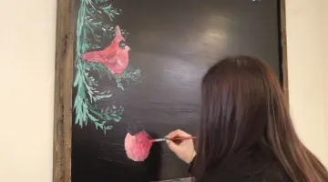 Maggie Russo uses chalk and paint to draw a colorful winter cardinal scene on a chalkboard.