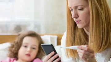 A woman looks at a thermometer while on a virtual visit with the pediatrician of her daughter, who sits in the background