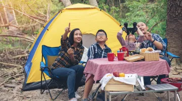 A family sits in front of a tent in the woods looking up at the sky and pointing at a bird.