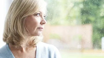 A woman stands near a window and looks sadly outside.
