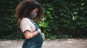 A pregnant African American woman poses outside while holding her stomach tenderly.