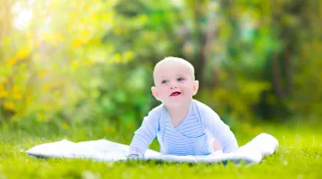 A happy baby boy does tummy time on a blanket outside in a green scene