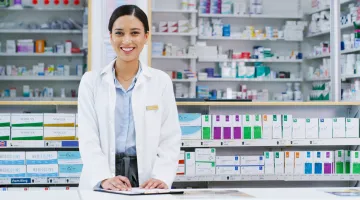 A smiling woman pharmacist stands at the counter with packages of medication behind her.