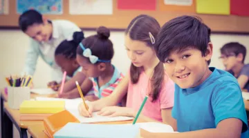 A young boy sitting at his desk in school writing on a piece of paper. Other classmates are doing similarly in the background.