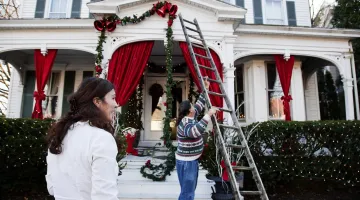 Man using a ladder to hang Christmas lights on his house