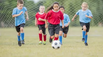 A girl in a red shirt and soccer uniform kicks a ball as four other players chase behind her in a youth soccer game.