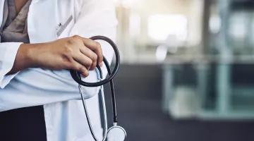 A close up view of a clinician in a white doctor's coat holding a stethoscope with a blurred office setting behind her