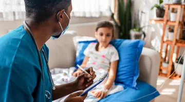 A male nurse writes on a clipboard as he speaks to a young girl patient, who is seated on a comfortable chair with an IV in her arm at her home.