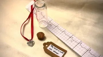 A metal heart pendant on a red ribbon, a cork, an empty class vial, a tag reading "my heart is with you, always," and a printed EKG heartbeat.