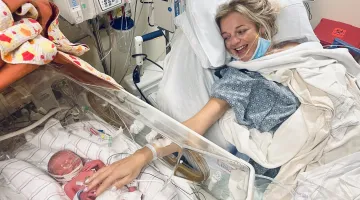 Baby Monroe lies in a NICU bed at South Shore Hospital shortly after birth, as mother Jillian reaches out to touch her, smiling, from her own hospital bed.