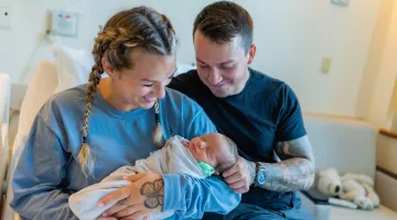 A mom smiles as she looks down at her newborn baby in her arms. Her partner touches the baby's head while he sits next to her.