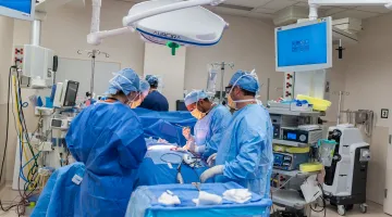 Three surgeons in blue scrubs, caps, and masks crowd around an operating table in an operating room at South Shore Hospital.